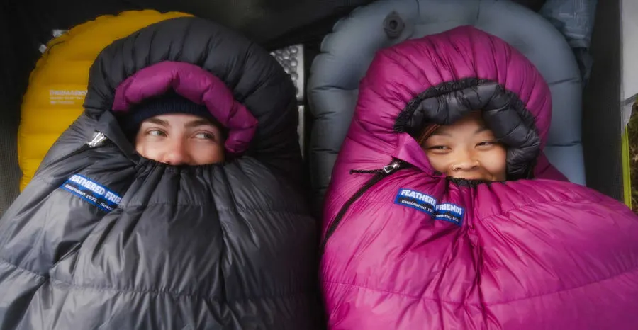 Two smiling campers, faces peeking out, are zipped into Feathered Friends down sleeping bags — one graphite grey, one magenta — each showing a blue Feathered Friends brand label at the collar.