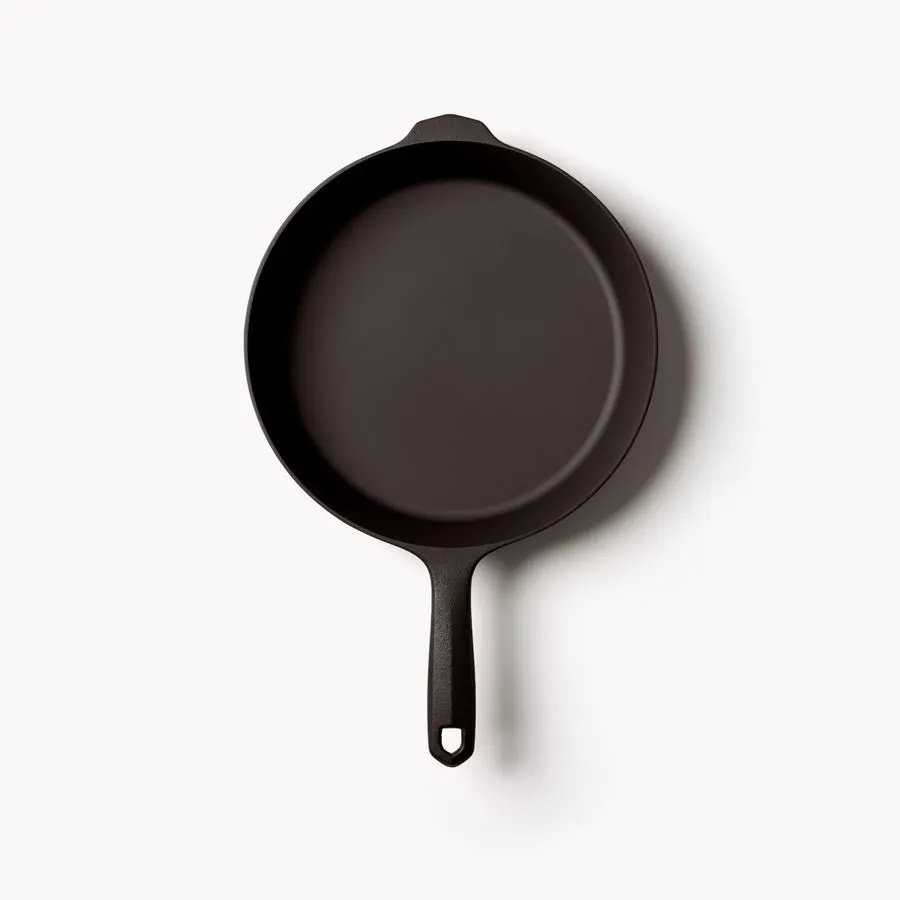 A dark seasoned cast iron skillet with a long handle and small pour spout, shot from directly overhead on a light gray background.
