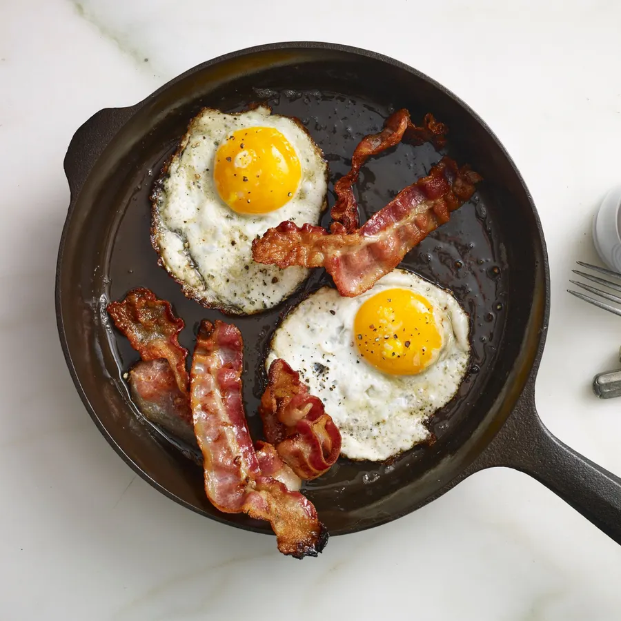 Overhead view of a dark cast iron skillet on a marble counter frying two sunny-side-up eggs and four strips of bacon, with a fork and salt shaker beside it.
