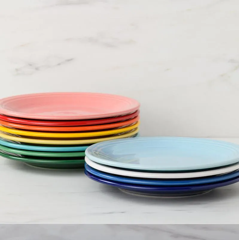 A stack of Fiesta dinner plates in coral, red, orange, yellow, green, and a second stack in sky blue, white, and cobalt, on a marble counter against a marble backsplash.