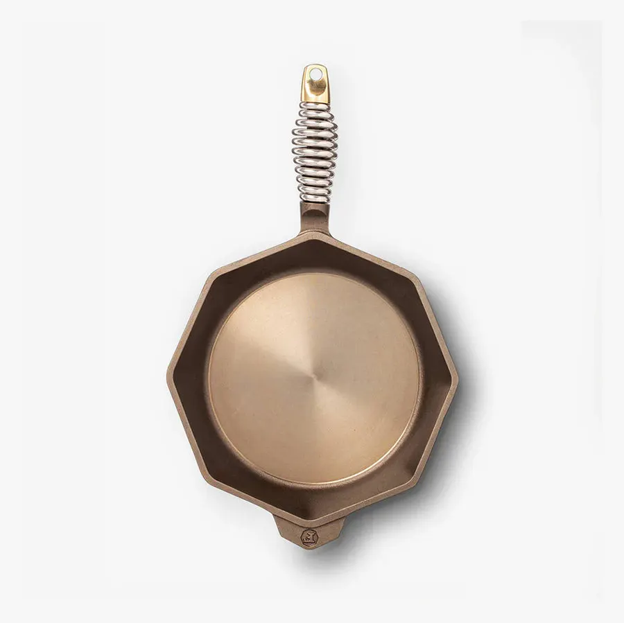 An octagonal cast iron skillet with a polished interior, dark seasoned rim, and a stainless coil spring handle with a brass ring, shot from directly overhead on white.