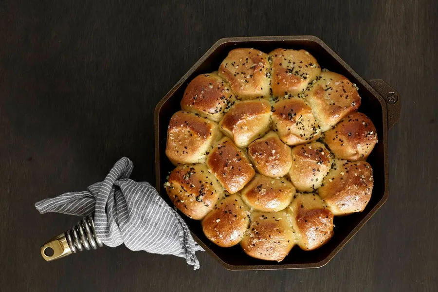 Overhead view of an octagonal cast iron skillet filled with a pull-apart pan of baked, seeded dinner rolls, with a striped cloth napkin looped through the coil handle, on a dark wood surface.
