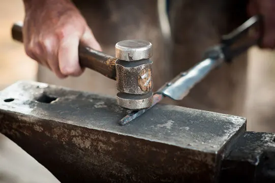 A blacksmith's hand strikes a hot, glowing garden-tool blade blank with a hammer on a steel anvil, sparks of scale visible on the metal.