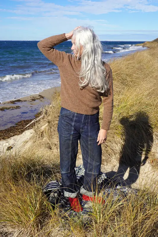A woman with long grey hair, wearing a brown wool sweater and indigo denim dungaree-style jeans, stands on a beach dune looking out at the ocean.