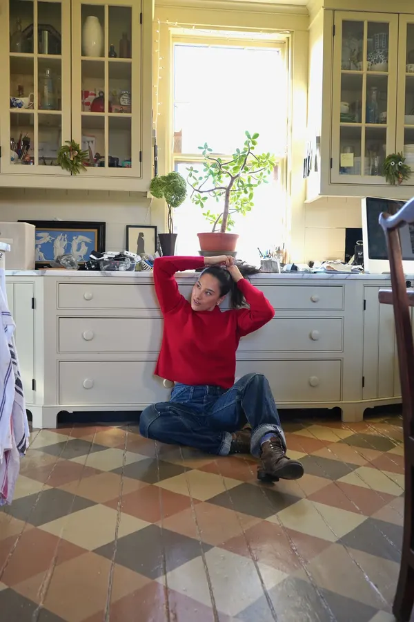 A woman in a red mock-neck sweater and denim jeans sits on a patterned tile kitchen floor, arms raised overhead, in a sunlit farmhouse kitchen.