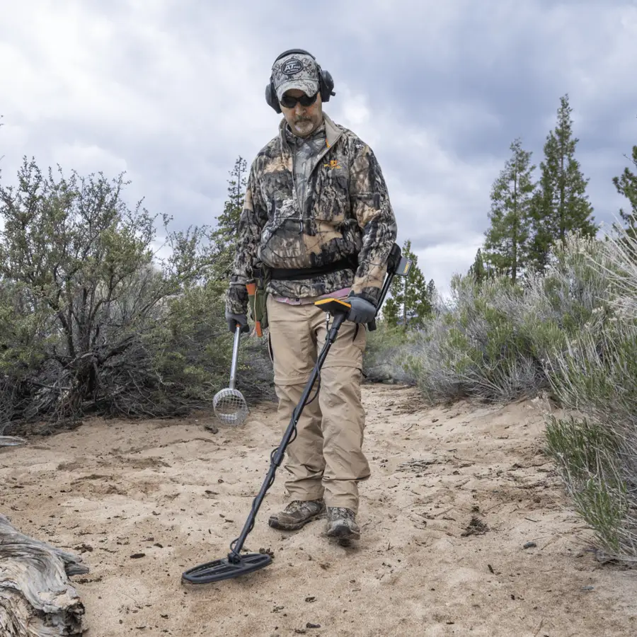 A person in camouflage jacket, cap, and headphones swings a black and yellow metal detector low over sandy desert ground scattered with sagebrush and pine trees.