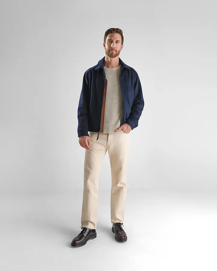 A man with a beard, wearing a navy Golden Bear zip jacket over a cream sweater, cream trousers, and black boots, standing full-length against a plain studio backdrop.
