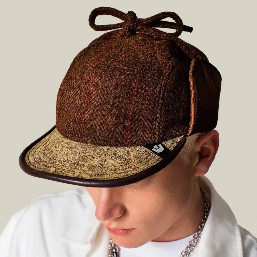 A young man in a white hoodie and chain necklace, looking down, wearing a brown herringbone tweed trapper-style cap with a tan suede brim and a corded chin tie knotted on top.
