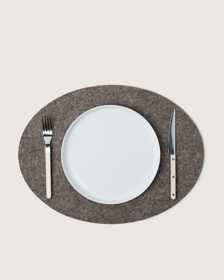 An oval taupe felt placemat set with a white plate, a fork and a knife, seen from above against a white background.