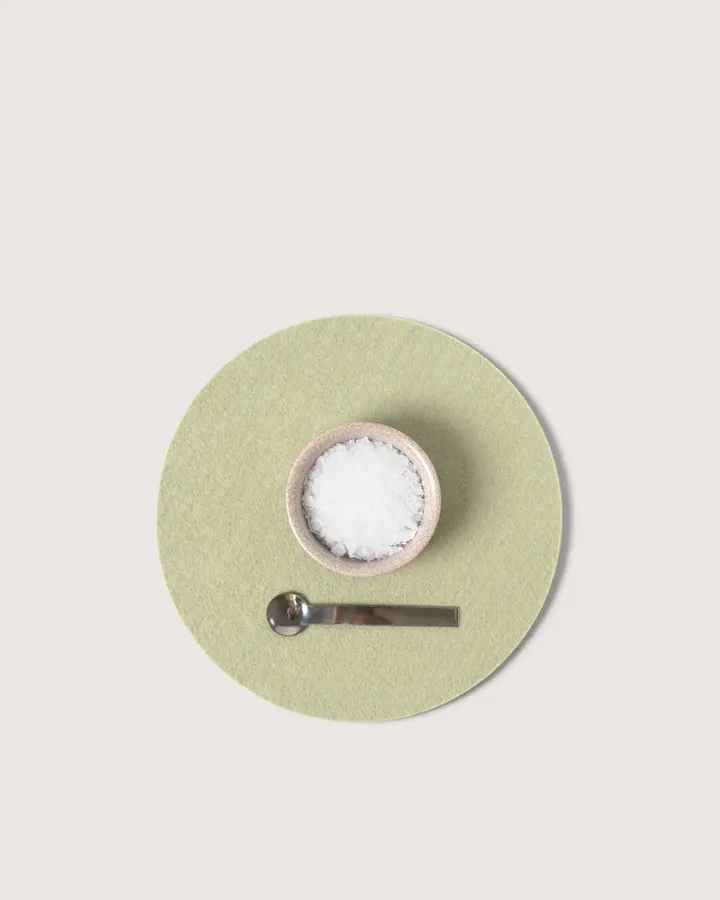 A round sage-green felt trivet holding a small ceramic bowl of salt and a mini spoon, seen from above against a white background.