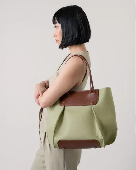 A woman in profile wearing a sleeveless beige jumpsuit carries a sage-green felt tote bag with brown leather trim over her forearm.