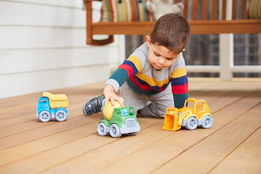 A toddler in a striped sweater kneeling on a wooden deck, reaching for a green Green Toys cement mixer truck, with a yellow dump truck and orange loader nearby.