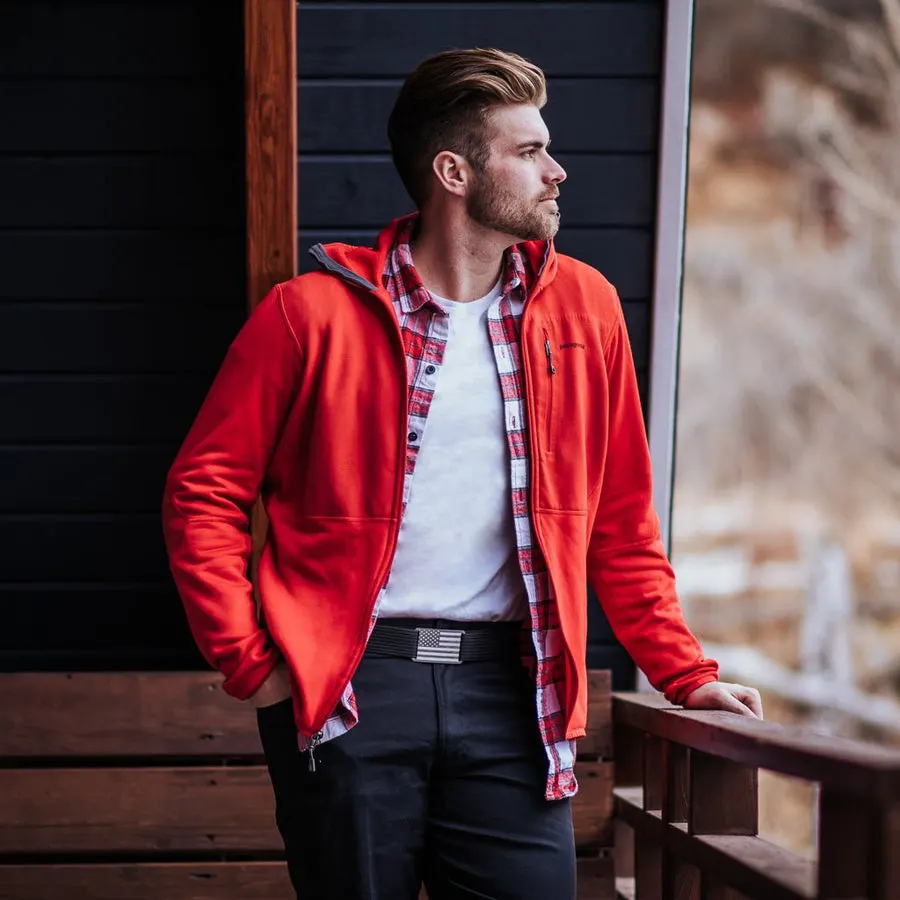 A man in a red zip-up jacket and flannel shirt stands on a wooden porch, hand resting on a railing, wearing dark pants with a black belt and a small American-flag buckle visible at the waist.