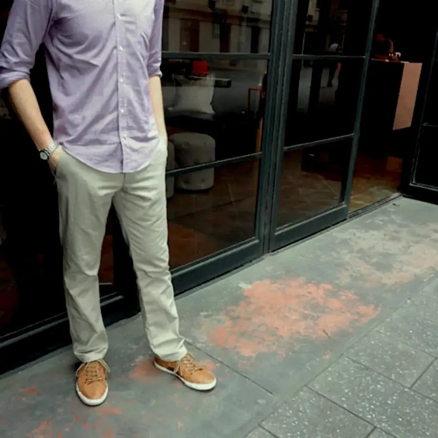 A man in a lavender button-down shirt and light khaki chinos with tan sneakers stands outside a storefront with glass doors, cropped from the shoulders down.