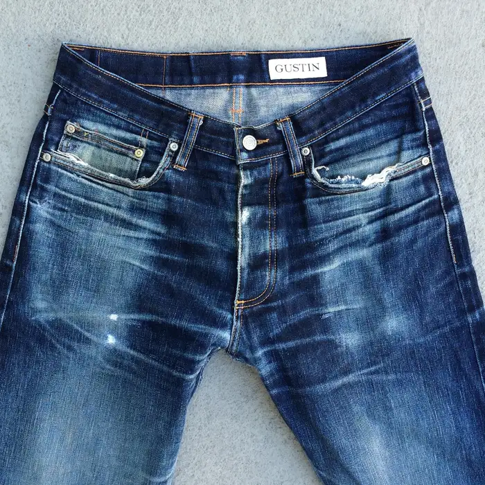 Close-up of dark indigo selvedge jeans laid flat, showing the waistband, coin pocket, button fly and natural fade whiskering at the hips.