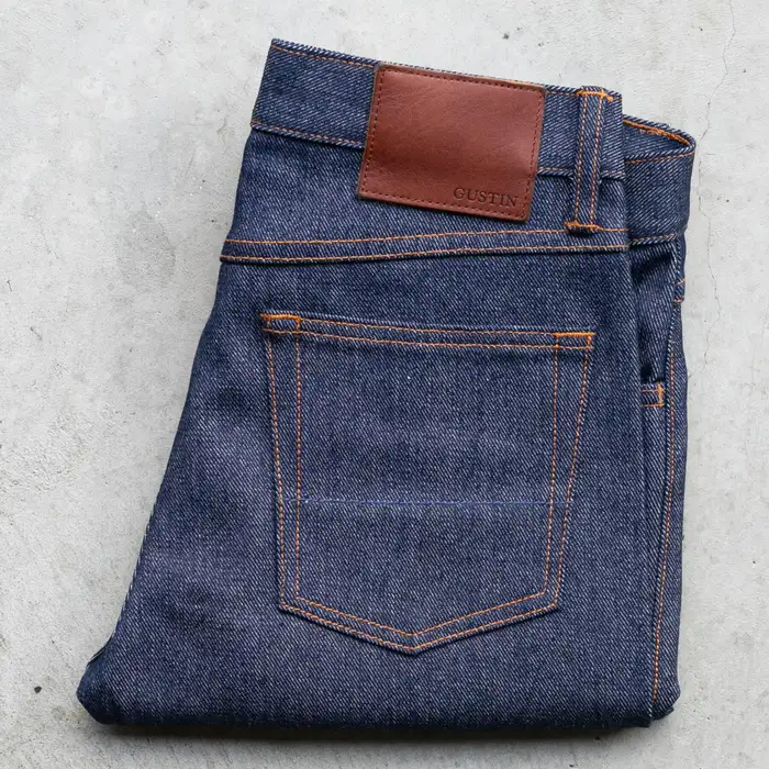 Dark raw denim jeans folded to show the back pocket with orange stitching and a brown leather Gustin patch, laid on a concrete surface.