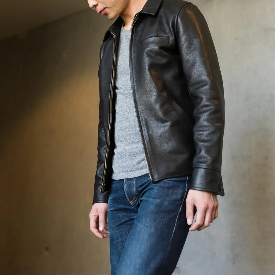 A man in a black leather jacket over a grey t-shirt and dark jeans, shown from the chin down against a concrete wall.