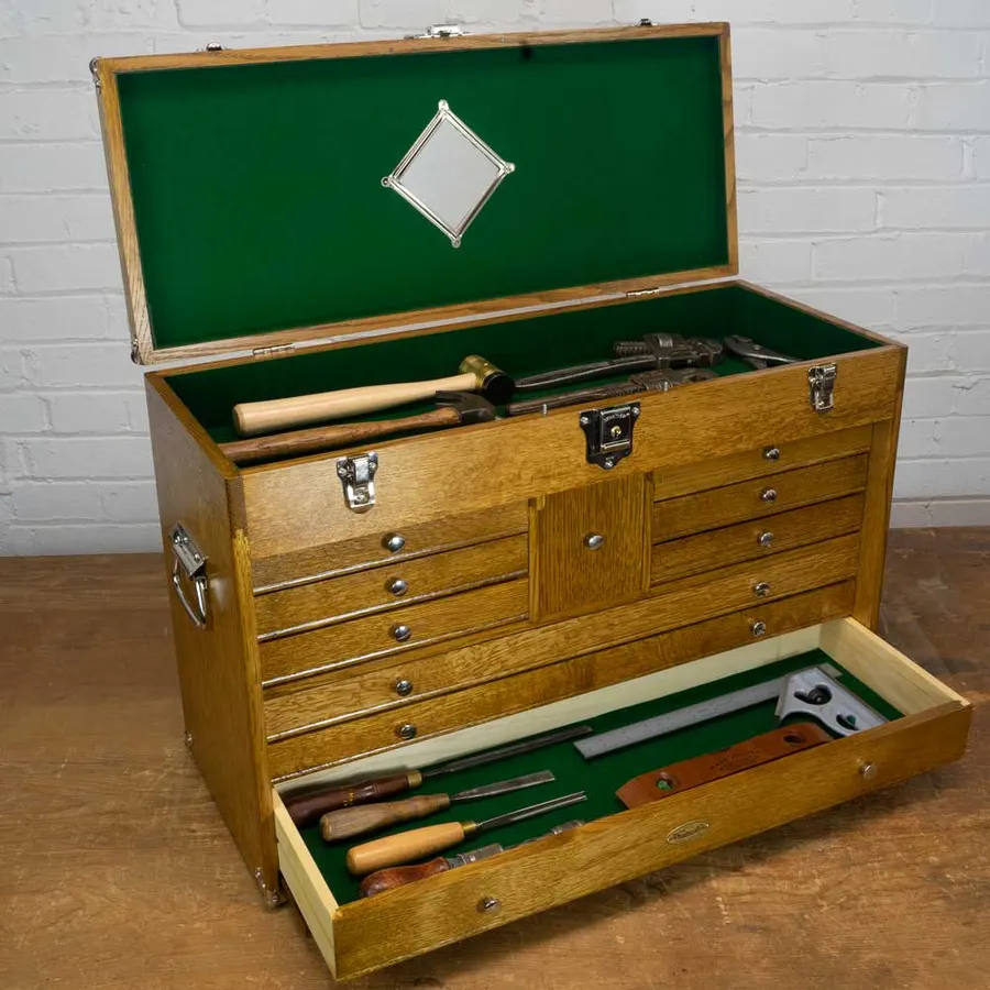 An open golden-oak tool chest with green felt-lined drawers and a mirror in the lid, its bottom drawer pulled out to show chisels, a hammer, and hand tools, against a white brick wall.