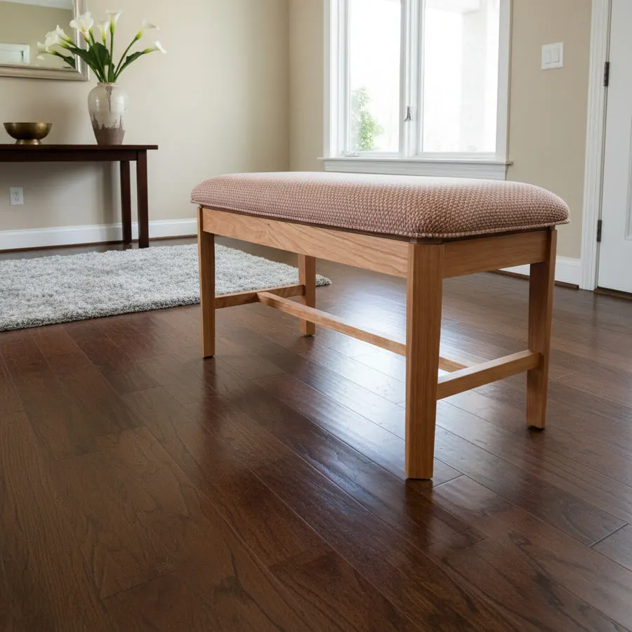 A wooden bench with a pale pink upholstered cushion top sits on a dark hardwood floor in a bright home entryway, with a rug, console table, and vase of flowers visible behind it.