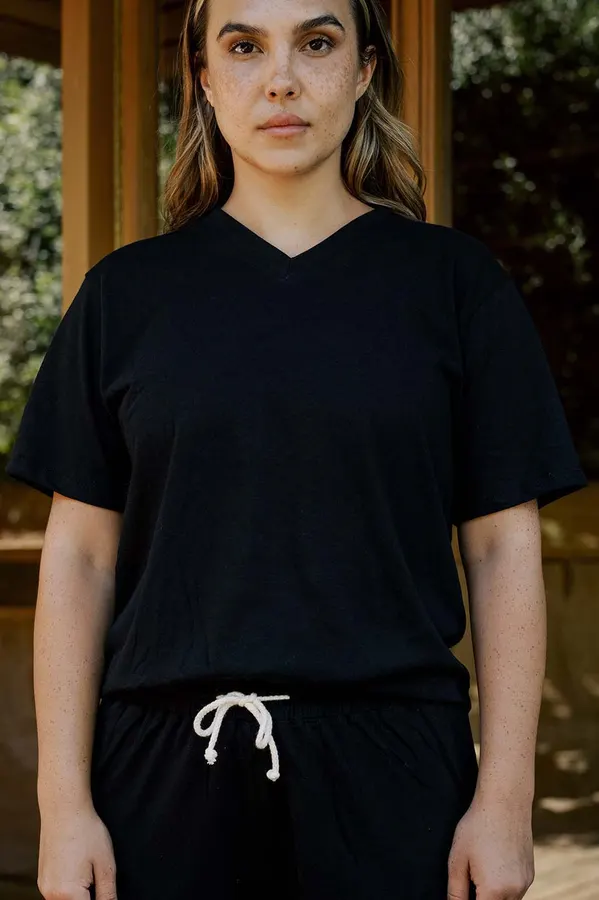 A woman with wavy hair wears a black V-neck organic cotton T-shirt and matching drawstring pants, photographed from the shoulders down to the waist outdoors against a wood-paneled backdrop.