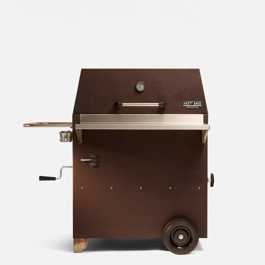 A Corten-steel charcoal grill on wheels with a slanted hinged lid, a built-in thermometer, a side crank handle, and a Hasty Bake nameplate, photographed against a plain light-grey background.
