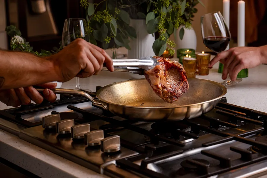 Two people's hands over a gas stovetop, one holding tongs lifting a seared steak from a stainless Heritage Steel fry pan, with wine glasses and candles softly lit in the background.