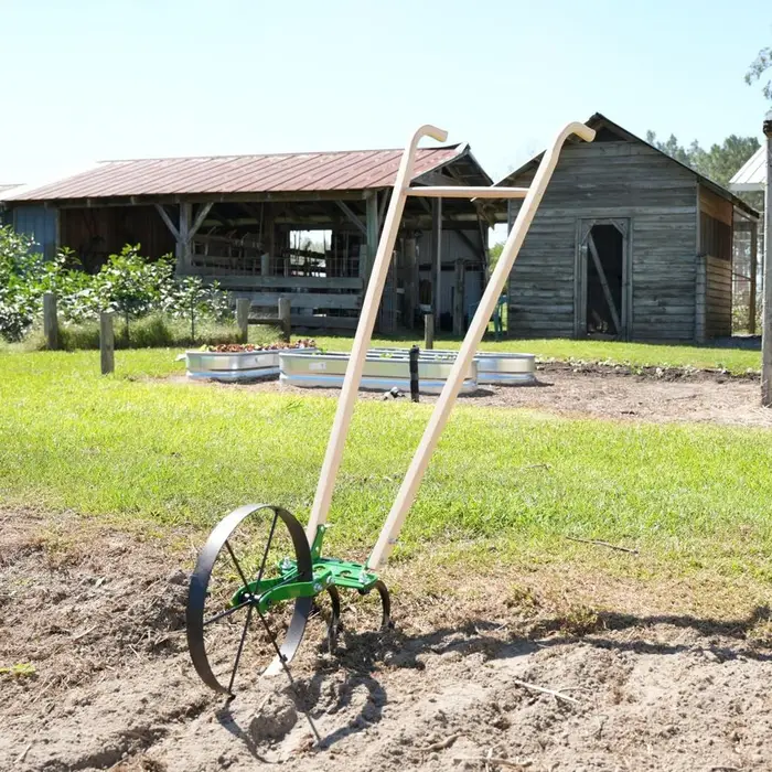 A green single wheel hoe with a large steel wheel and wooden handles stands upright in a garden bed, with raised metal planting beds and wooden farm outbuildings in the background.
