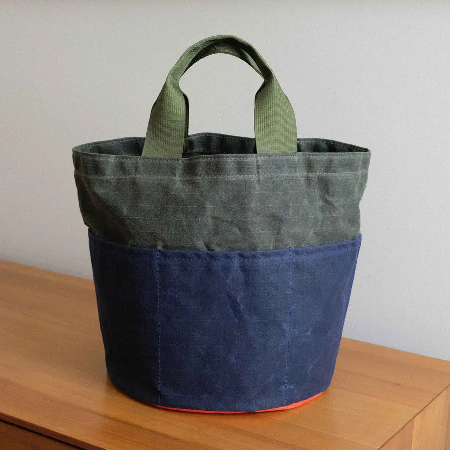 A waxed-canvas bucket bag with olive-green upper panel, navy lower panel, an orange trim strip at the base, and webbing handles, sitting on a wooden dresser against a plain wall.