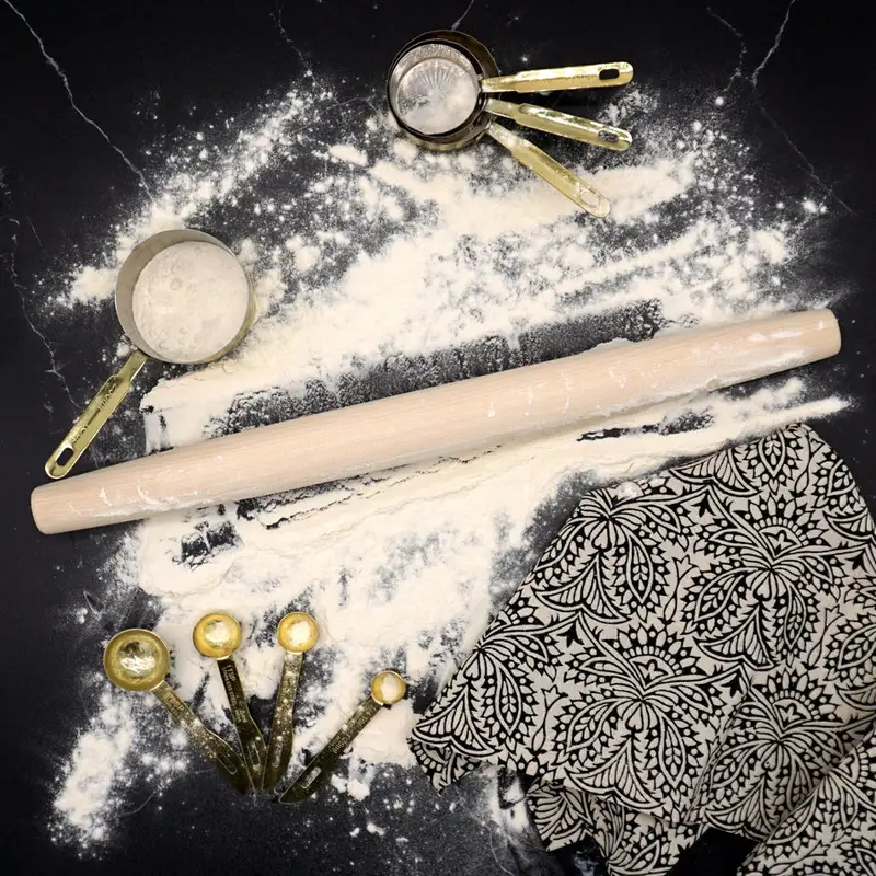 A pale wood rolling pin lying across a black countertop dusted with flour, surrounded by vintage brass measuring cups, measuring spoons, and a black-and-white patterned cloth.