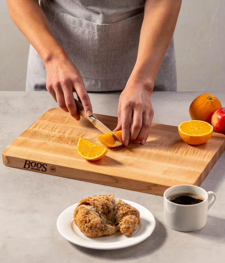 A person in a grey apron slicing an orange wedge with a paring knife on a Boos Block maple cutting board, with a croissant on a plate and a cup of coffee in the foreground.