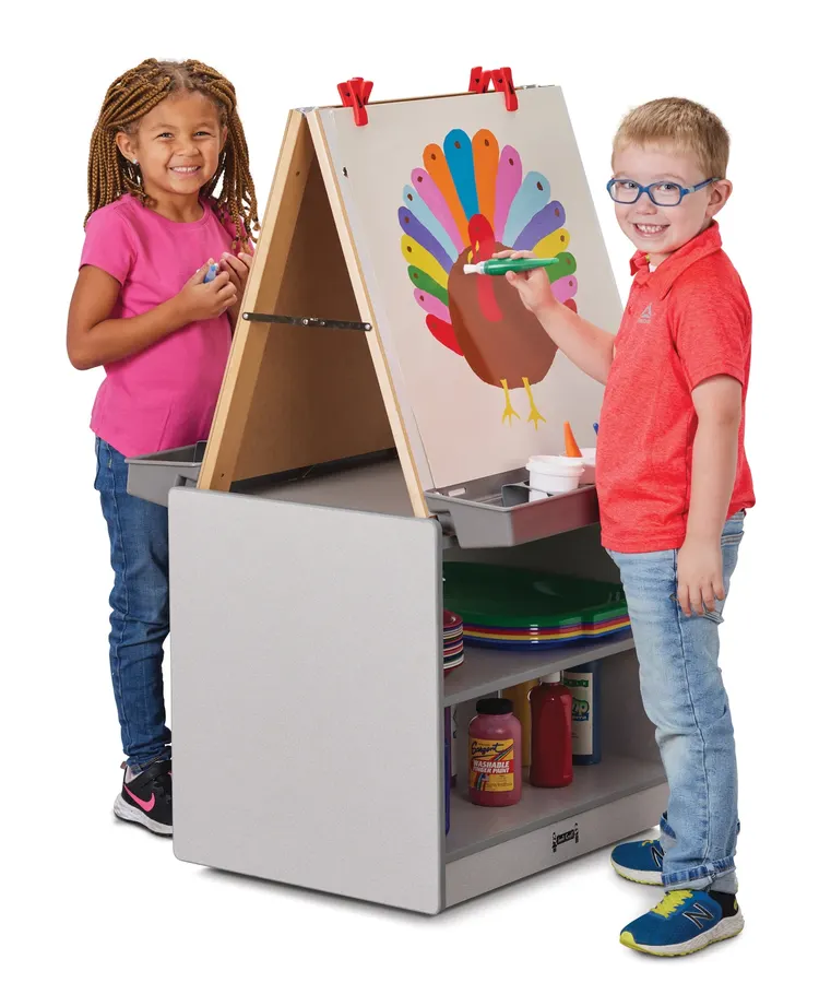 Two children in school clothes stand at a wooden A-frame easel with a storage cubby base, one holding a marker to a colorful turkey drawing clipped to the easel.