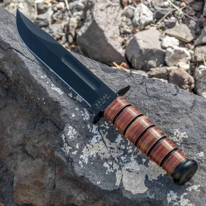 A classic KA-BAR fighting knife with a black blade and brown leather-washer handle, resting diagonally across a large grey rock outdoors.