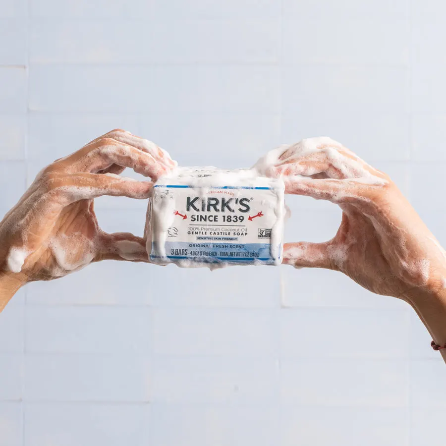 Two soap-lathered hands holding up a 3-bar pack of Kirk's Original Fresh Scent Castile Soap against a light blue tiled wall.