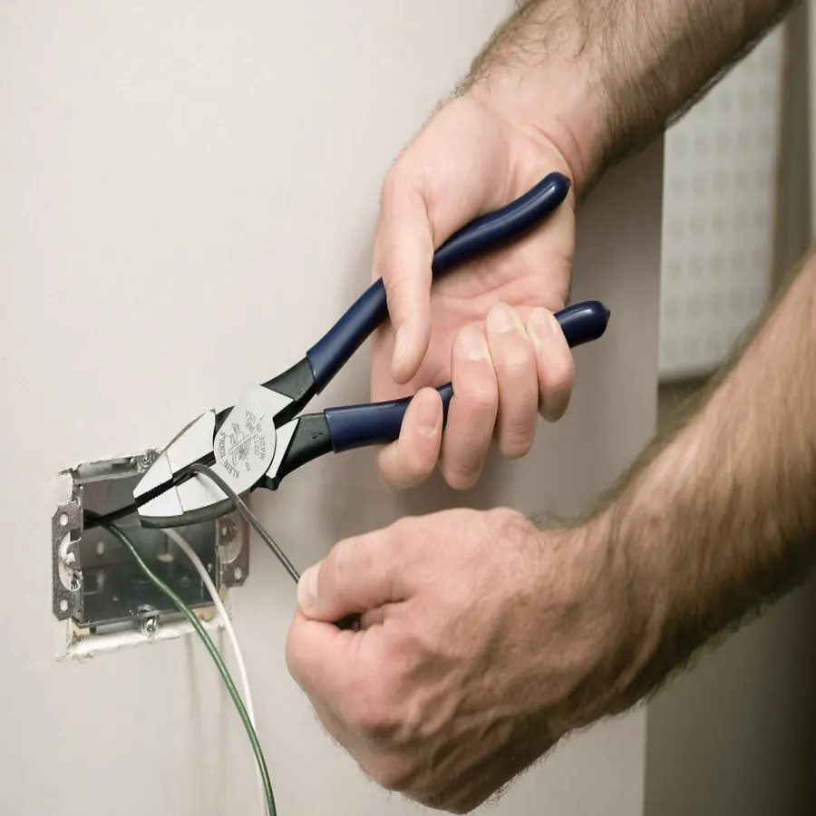 A close-up of two hands using navy-handled Klein Tools lineman's pliers to work on wiring inside an open electrical wall box.