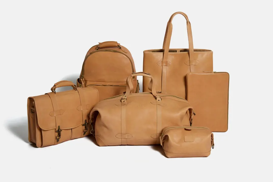 A grouped studio shot of six tan leather goods standing together on a plain background: a backpack, a rounded-top tote, a duffel bag, a dopp kit, a messenger-style briefcase, and a flat folio.