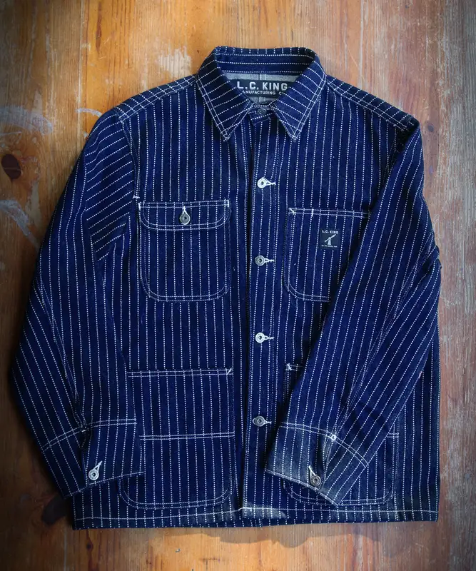 A wabash-stripe (navy with white pinstripe) denim chore coat laid flat on a wood plank floor, showing four patch pockets, metal buttons, and an L.C. King Manufacturing Co. neck label.