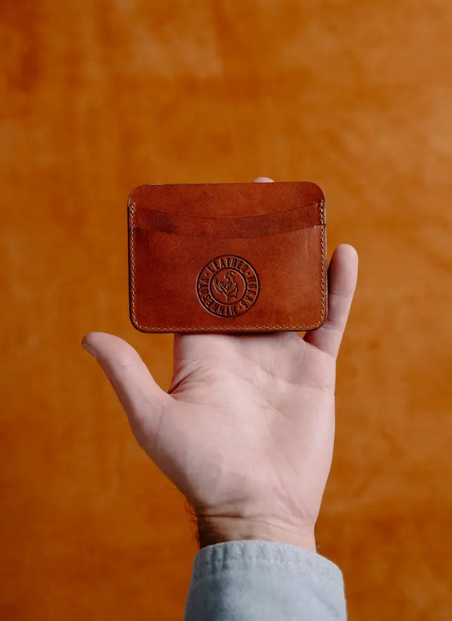 A hand holds up a small brown leather card wallet stamped with the Leather Works Minnesota circular logo, shot against a rich orange textured leather background.