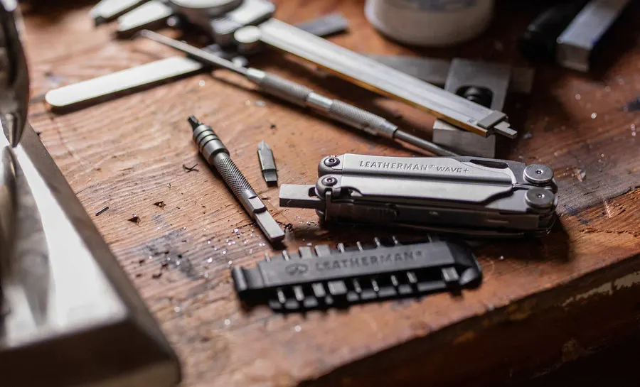 A closed Leatherman Wave+ multi-tool and its bit driver extension resting on a scarred wooden workbench among files, calipers, and a scriber.