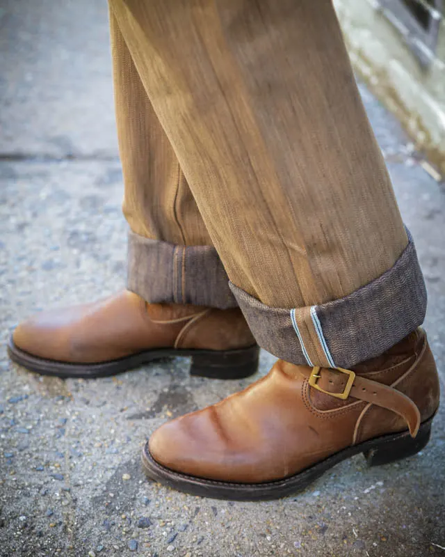 A person's cuffed olive-brown trousers and brown leather buckle-strap boots, shown from the knee down while standing on a concrete city sidewalk.