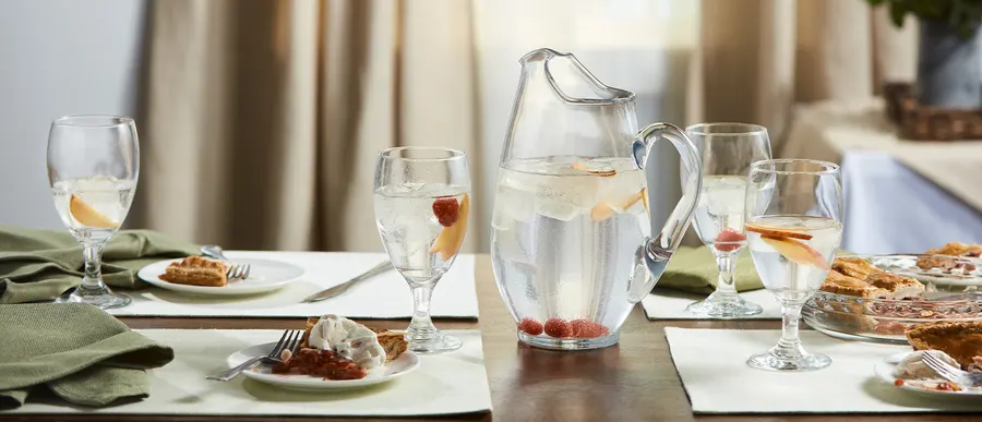 A set dining table with a glass pitcher of fruit-infused water, wine goblets filled with water, and plates of waffles and pie, framed by sheer curtains in soft daylight.