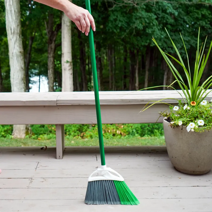 A hand holding a green-handled angled broom, sweeping a wood-look deck beside a stone planter with white flowers and a tree line in the background.