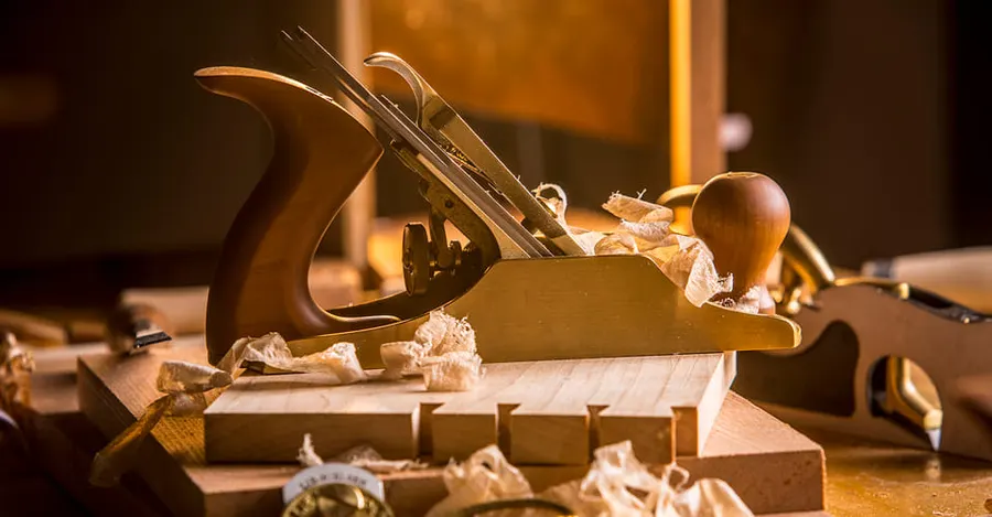 A bronze Lie-Nielsen No. 4 smooth plane resting on a dovetailed wooden workpiece surrounded by curled wood shavings, warmly lit, with a second plane blurred in the background.