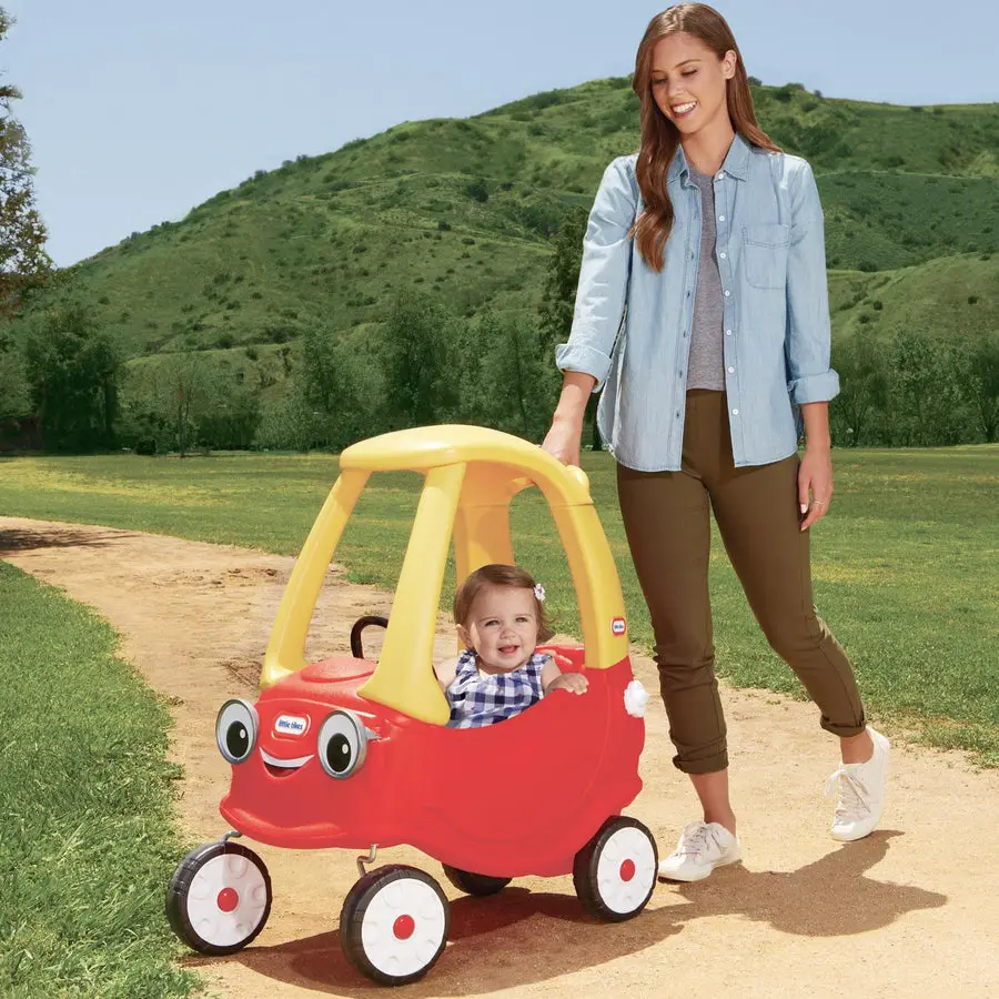 A woman in a denim shirt walking a smiling toddler seated in a red-and-yellow Little Tikes Cozy Coupe ride-on car along a dirt path with green hills behind them.