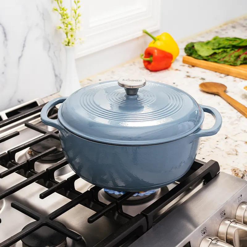 A slate-blue enameled cast iron Dutch oven with the lid on, sitting on a lit gas stovetop burner in a kitchen with marble backsplash, bell peppers, and greens in the background.