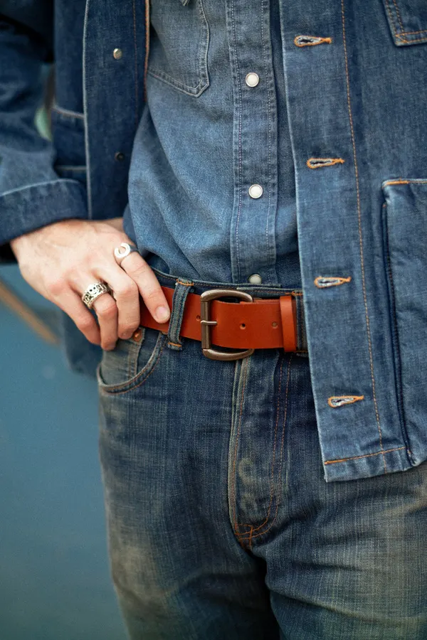 Close crop of a man's waist wearing a tan leather belt with an antique brass buckle over dark selvedge denim jeans and a chambray jacket, one hand tucked into the pocket.