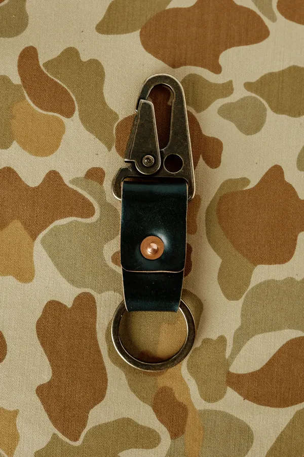 A black leather key clip with a brass swivel hook and copper rivet, attached to a keyring, laid flat on a camouflage fabric background.