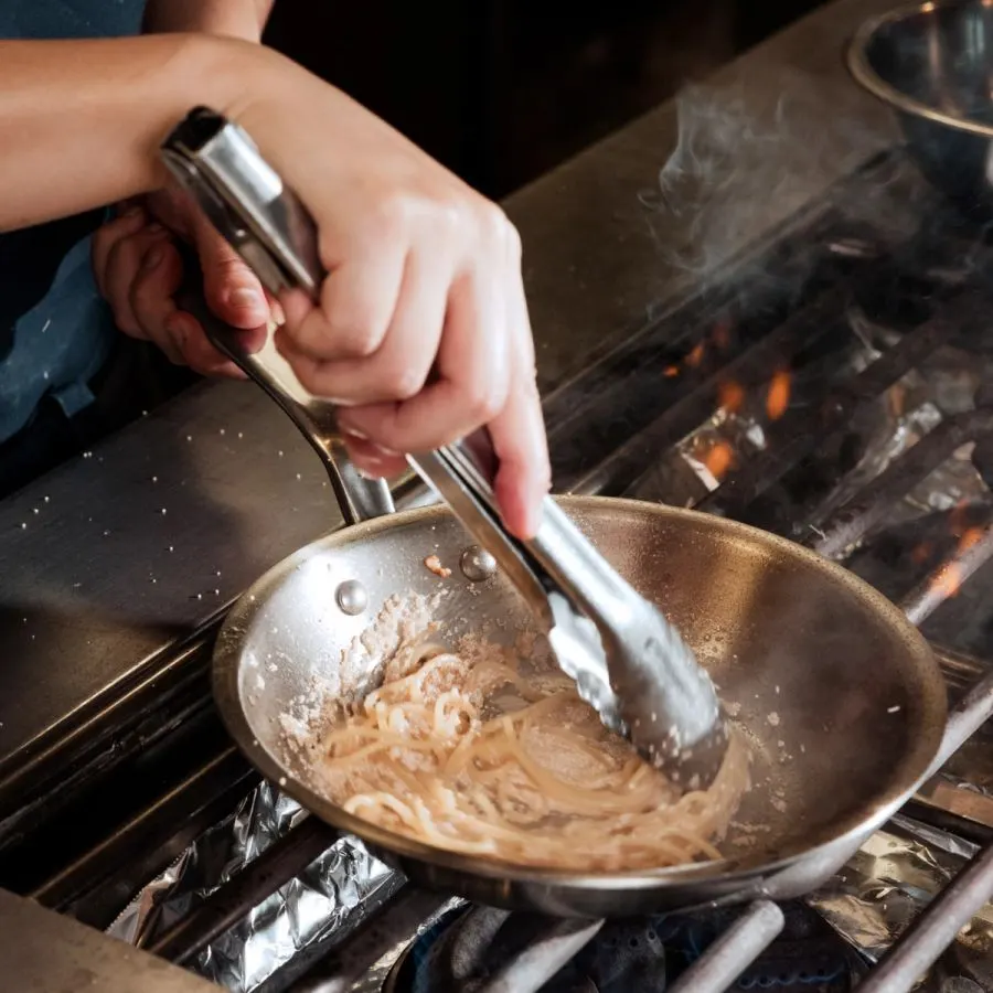 A pair of hands using tongs to toss pasta in a stainless steel frying pan over an open flame on a professional grill, with steam rising.