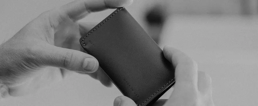 Black-and-white close-up of two hands holding open a slim black leather bifold wallet stamped 'Made in USA', with a blurred room in the background.