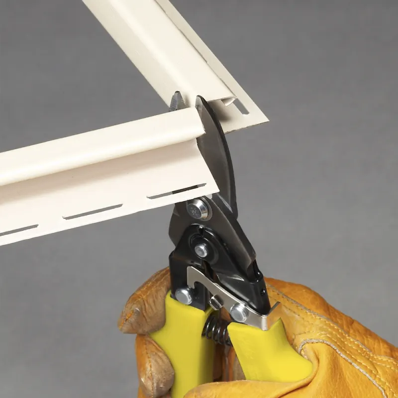 A gloved hand using black-and-yellow Malco Max2000 aviation snips to cut a piece of white vinyl siding trim at a corner joint.