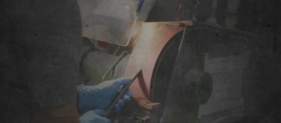 A worker in blue gloves and a face shield grinding the edge of a metal tool blade against a bench grinder, sparks flying, rendered with a dark grungy photo treatment.